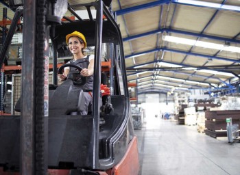 Professional female industrial driver operating forklift machine in factory's hall.