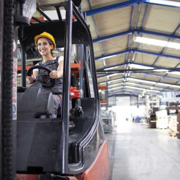 Professional female industrial driver operating forklift machine in factory's hall.
